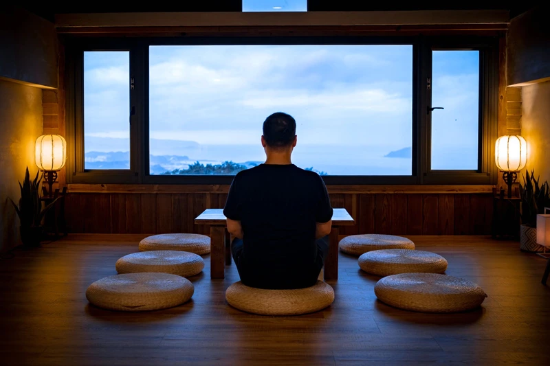 man with his back to the camera in the lotus position looking at the horizon through a large window